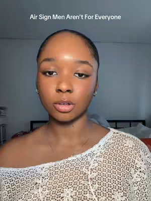 A medium shot shows a young woman with dark skin against a neutral background. She has smooth skin, light makeup, and her dark hair is pulled back. She’s wearing a white, lace-like top and small silver earrings. The text overlay reads 'Air Sign Men Aren’t For Everyone.' The shot appears to be taken indoors, with a bed and part of a room visible in the background.