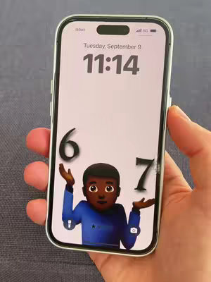 A hand holding an iPhone displaying a lock screen with the time and a cartoon character emoji with black skin offering the numbers '6' and '7'.