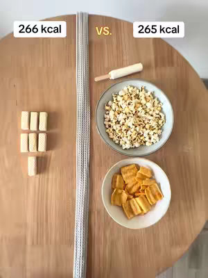 An overhead shot displays a wooden table divided into sections. The left side features five rectangular wafer cookies and the text '266 kcal' in a white box. A long, thin strand of what appears to be noodles or pasta runs vertically through the center. The right side includes a popsicle on a stick, a bowl of popcorn, and a bowl of what appears to be fried plantains or chips with the text '265 kcal' in a white box. The items are arranged to visually represent different calorie counts.