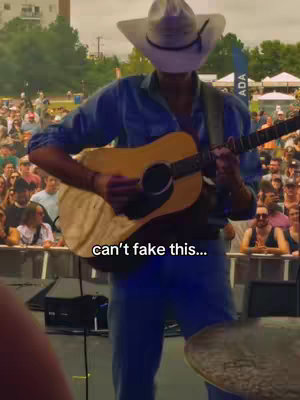 A man wearing a cowboy hat is playing a guitar on stage in front of a crowd. The image is shot from a low angle. Text overlay reads 'can't fake this...'.
