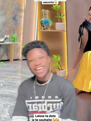 A smiling young man is the central focus against a blurred interior background. He's wearing a t-shirt with text and a silver chain. The background includes a decorative shelf with plants and a glimpse of a woman. Overlay text at the top and bottom of the image adds context and a call to action.