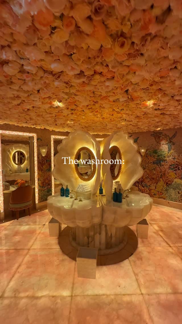 The image presents a lavish, ornate washroom in Dubai. The focal point is a double sink that is shaped like a giant, open seashell, with gold-rimmed mirrors and fixtures. The ceiling is covered in a dense array of peach-colored artificial flowers. Walls are adorned with floral mosaics, and soft lighting enhances the opulent ambiance. The floor features a pink-toned marble tile, and the whole scene feels luxurious and highly stylized.