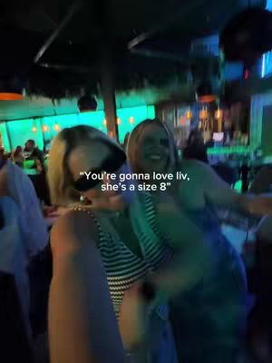 Two women are posing for a selfie in a bar or club setting, with a blurred background of lights and people. The women are close together, with one in the foreground wearing sunglasses and the other smiling. The lighting is dim, typical of an indoor social setting.