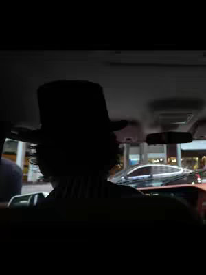 The video cover image shows a person from behind, seated inside a vehicle, facing forward. The person, silhouetted against the car's interior, wears a top hat. The ambient light casts dark shadows, obscuring the details of their face and attire. Through the windshield, a blurred view of the city lights and other vehicles can be seen.
