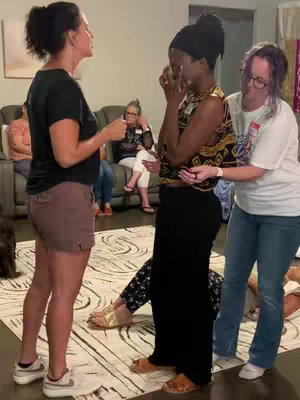 The image shows three women. One woman is in a black top and pants. She has a black head covering and is holding her face in her hands. A second woman in a black top and brown shorts is talking to the first woman, with her arm outstretched in a supporting manner. A third woman in a white t-shirt and glasses has her arm around the first woman. There are other people in the background, including some on a couch. The overall color scheme is warm and inviting.