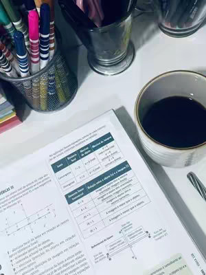 A top-down shot of a study space features an open textbook with detailed diagrams and text in Portuguese. Beside the book sit a white mug filled with black coffee and a pen. Colorful markers and pens are arranged in wire and glass containers. The scene is illuminated with warm, natural light, creating a cozy study atmosphere.