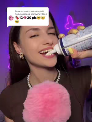A woman is shown in a close-up shot, holding a container of what appears to be whipped cream and putting it in her mouth. She is smiling and has earrings. Behind her is a blurred background with a neon sign and a microphone.