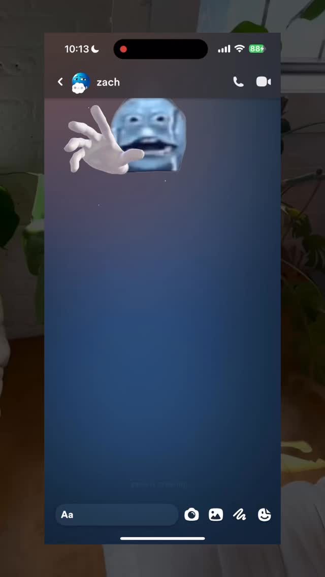 A phone display showing a messaging app screen with a funny AI-generated face overlay. The background is a blurred view of a room with plants.