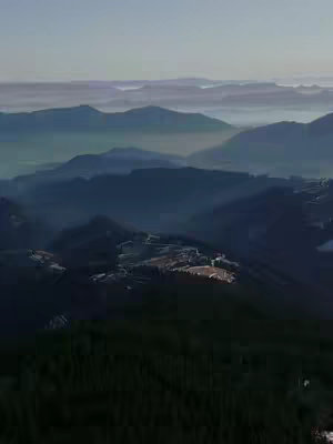 An aerial view of a mountain range at dusk or dawn, with layers of peaks fading into the misty distance. The sky has a gradient of colors, transitioning from a light blue to a slightly darker shade, indicating either sunrise or sunset. The mountains in the foreground have visible surface details, while those in the background are softened by atmospheric perspective.