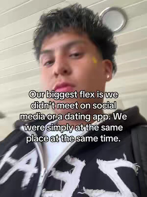 A selfie-style shot features a young man with dark hair and a neutral expression. He's wearing a black hoodie with white graphic elements on it, and the top portion of his body fills most of the frame. The background shows a white ceiling and a light fixture, with text overlayed describing the genesis of his relationship.