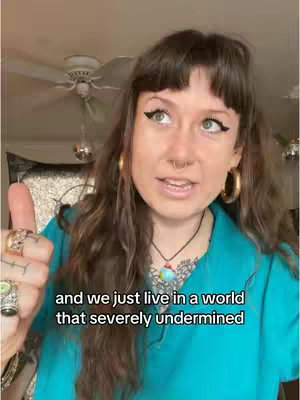 A woman with long brown hair and bangs is giving a thumbs-up. She is wearing a turquoise shirt, gold hoop earrings, and multiple rings. She has a nose ring and eyeliner. The background includes a ceiling fan and hints of interior decor.