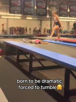 The image shows an indoor setting, likely a gymnasium, with two cheerleaders on a blue mat. One is lying down with arms outstretched. The other is standing with one leg slightly elevated, appearing to be preparing to perform a move. There is text overlayed, a portion stating 'born to be dramatic, forced to tumble'.