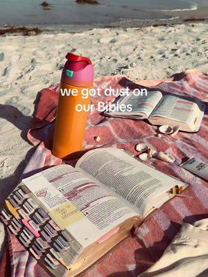 A beach scene with books and a water bottle, suggesting a place for quiet reflection and reading. A bottle, open Bible sits on a blanket, with sand and ocean visible.