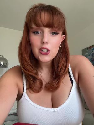 The cover image features a woman in a close-up, centered shot. She has fair skin, red hair styled with bangs, and is wearing minimal makeup with bold eyeliner and lip color. She is wearing a white tank top, her hands are out of frame, with a light-colored wall and a piece of furniture in the background. The overall composition is well-lit, focusing directly on the woman's face.