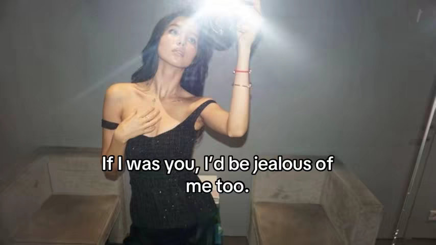 A young woman poses for a selfie indoors, holding a light source above her head with her right hand. The lighting creates a bright flare, and the background is a blurred, neutral gray. She is wearing a black, sparkly top and has long, dark hair. Text overlayed at the bottom states: "If I was you, I'd be jealous of me too."