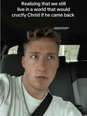 A young man with short, light-colored hair and a white t-shirt is seated inside a car. The background appears to be the interior of the vehicle, with a window showing a glimpse of an outdoor setting. The man is looking towards the left of the screen, with a contemplative expression. Above the image are overlaid white text that reads, Realizing that we still live in a world that would crucify Christ if he came back.