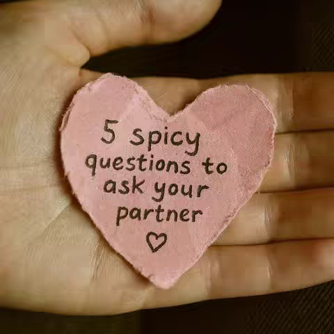A close-up shot of a light hand holding a heart-shaped piece of pink paper. Text in black is handwritten on the paper: "5 spicy questions to ask your partner" with a small heart symbol below.