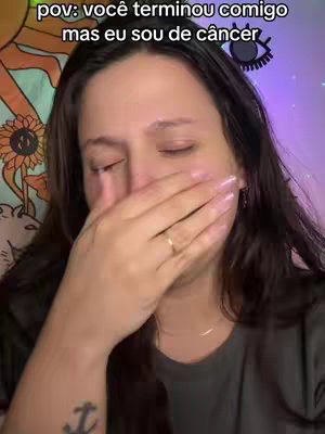 A woman with dark hair is shown crying with one hand covering her mouth. She has a faint gold ring on her wedding finger. The background is blurred, but includes a wall with a tapestry and a purple light. There is text overlayed at the top that reads: “pov: você terminou comigo mas eu sou de câncer,” which translates to 'pov: you broke up with me, but I am cancer.'