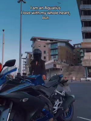 A person wearing a black helmet and jacket is standing next to a dark blue motorcycle with a city background. The sky is clear with some buildings in the background. There appears to be some text at the top of the image.