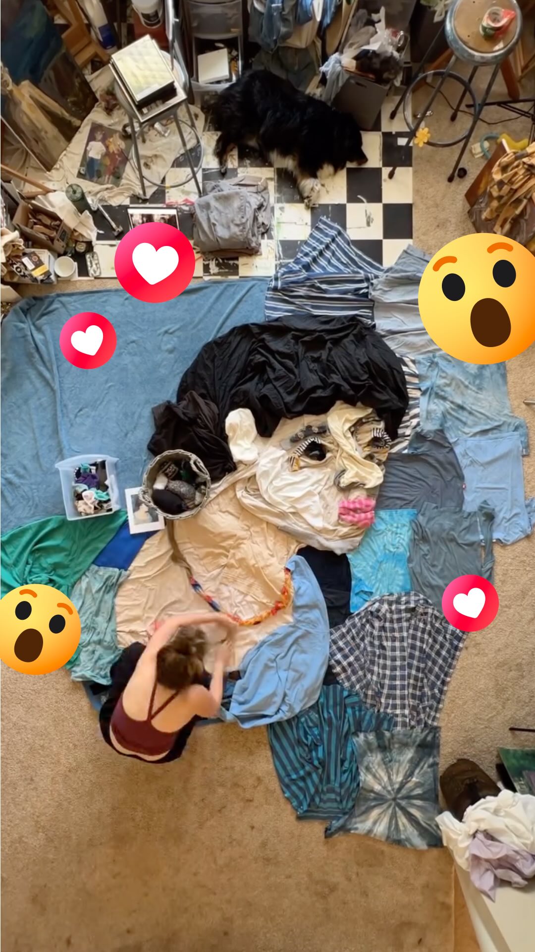 An overhead shot shows a woman creating a fabric portrait on the floor using various colored and patterned clothing items. The face, made of folded fabrics, is placed against a large, light blue sheet. A dog is visible in the back of the messy room. Facebook reaction emojis are scattered around.