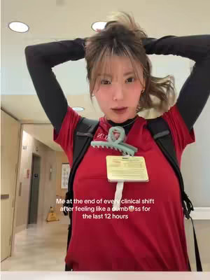 A young woman with light brown hair pulled up in a bun, wearing a red scrub top and black undershirt, is shown from the chest up. She's holding her hands up to adjust her hair. A backpack and ID badge are visible. The background shows a hallway, including a wall and fluorescent lights.