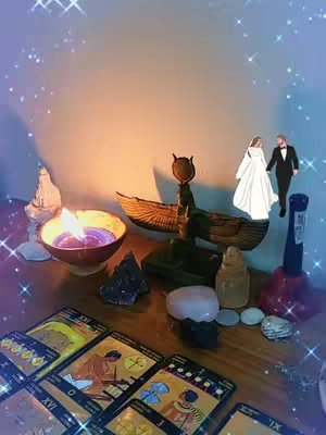 The image is of a tarot card reading setup with several cards fanned out on a wooden table. A lit candle in a decorative holder is in the foreground, casting a warm glow. Various crystals, figurines, and a bird-shaped object sit on the table, and a miniature bride and groom are positioned in the background. Stars and sparkles are added to the top of the layout to visually enhance the scene.