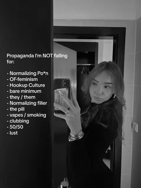 A grayscale selfie-style image features a young woman, positioned slightly off-center, looking towards the right. She's holding a phone in front of her, likely taking a selfie or recording a video, and looking off-camera. The setting appears to be indoors, possibly a hotel room or apartment, with a partially visible black door and a mirror in the background. Text overlayed on the left side of the image lists phrases, suggesting a list of things the individual is 'not falling for'.