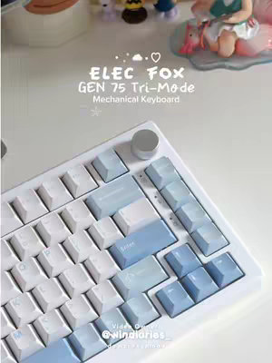 A top-down shot showcases a white and light-blue mechanical keyboard on a white surface. Above the keyboard, various small decorative items and figurines are slightly out of focus. The image is well-lit, with cool tones dominating and minimal shadows, giving a clean and inviting feel.