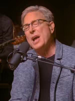 The image shows a man with gray hair and eyeglasses singing into a microphone. He is wearing a blue blazer over a black shirt. The background is dark, and another person playing a guitar is partially visible.