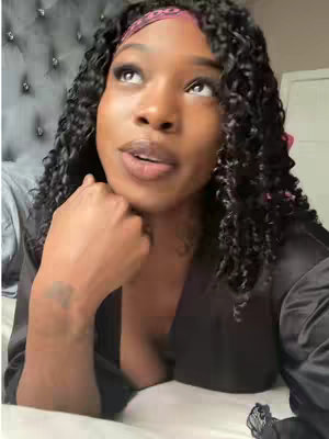 A medium shot shows a Black woman lying on a bed, gazing upwards. She wears a black satin robe over what appears to be a matching or contrasting top. Her dark hair cascades down, and a pink headband holds it back. Her expression is contemplative. The background shows a tufted headboard and a glimpse of a white door.