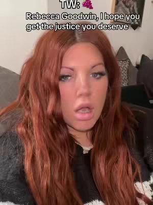 A woman with long, red hair is looking directly at the camera with an open mouth, as though speaking. She has fair skin, rosy cheeks, and is wearing a dark top. The background is slightly blurred, suggesting she may be in a living room.