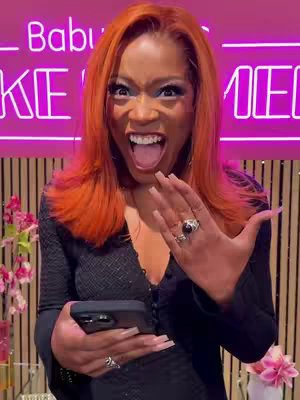 A person with bright orange hair and dramatic makeup is reacting with open-mouthed surprise, holding a smartphone and displaying their hand with eye-catching rings. The background contains a neon sign and wooden vertical paneling.