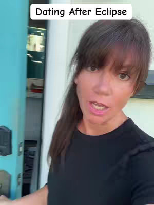 A woman with brown hair wearing a black shirt stands in front of a blue door and speaks directly to the camera, with text overlayed above reading "Dating After Eclipse"