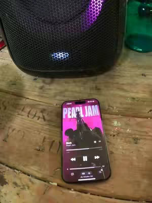 The image shows a close-up of a wooden surface with a smartphone and a speaker, with a green water bottle in the background.