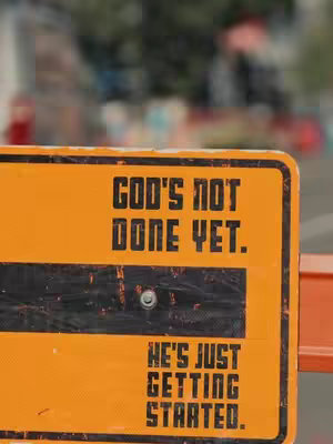 The image is a close-up shot of a yellow and black road sign. The sign has bold, black text that reads "GOD'S NOT DONE YET. HE'S JUST GETTING STARTED." Behind the sign, the background is blurred, suggesting a street scene with blurred details.