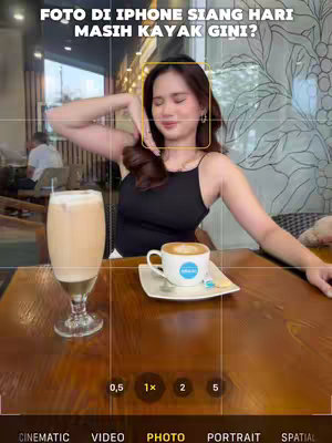 A photo taken during the day shows a woman in a cafe holding her hand up and smiling. A coffee drink and coffee cup is on the table in front of her on a wooden table.  A camera focus box is around her face.