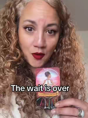 A woman with curly brown hair, fair skin, and dark red lipstick holds a tarot card in front of her chest. The card features a woman in a pink and blue bubble. The woman in the image is smiling with her eyes looking towards the camera with a slight tilt in her head. The text "The wait is over" is superimposed on the image in white letters.