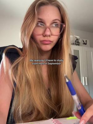 A young woman with long blonde hair and glasses looks at the camera with a concerned expression while holding a pen and notebook. Text overlaid on the image reads, 'Me realizing I'll have to study even more in September'.