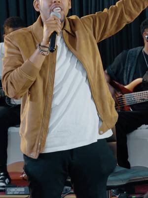 The cover image depicts a man singing into a microphone during a performance. He is wearing a light brown jacket over a white t-shirt and black pants and bracelets . Other band members can be seen blurred in the background.
