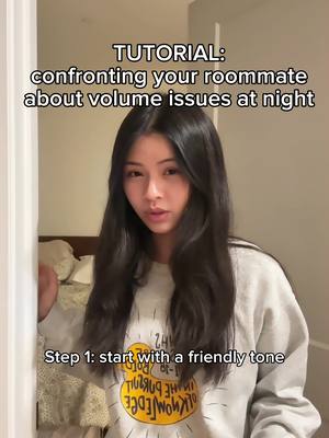A young woman with long, dark hair is centered in a video frame, standing indoors. Text overlays describe this as a tutorial on how to address volume issues with a roommate. She wears a gray sweater with a yellow and black graphic. Behind her, a bed can be partially seen, suggesting the setting.