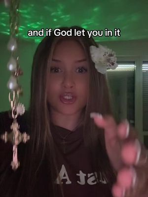A young woman with long brown hair is facing the camera with an open mouth, as if she is speaking. She has a white flower in her hair and is holding rosary beads towards the camera with her right hand. A hook, "and if God let you in it" in placed above her head.