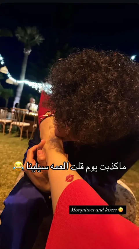 An intimate shot shows a person kissing another person's arm, adorned with a lipstick tattoo. The scene takes place at an outdoor, night setting with subtle lighting that creates a warm atmosphere.