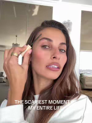 A woman is holding a makeup brush to her eyebrow. The image background is slightly blurred but reveals a large mirror and a door. There's a text overlay on the bottom of the image.