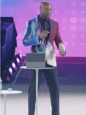 A man in a colorful blazer stands on a stage in front of a purple backdrop, holding a microphone. A small table with a tablet sits in front of him.