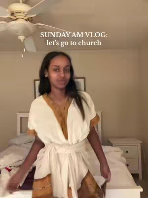 A woman is standing inside a room near a bed. She is wearing a dress and smiling towards the camera. The room has a bed, a ceiling fan, and a picture on the wall.