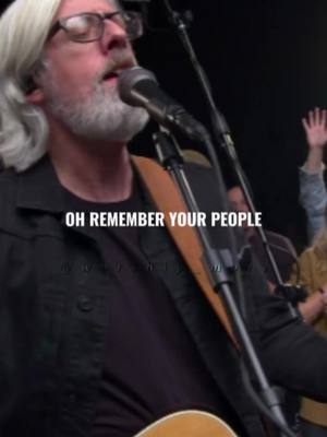 A man with gray hair and a beard singing into a microphone with his eyes closed, wearing glasses and a black jacket. The text "OH REMEMBER YOUR PEOPLE" is overlaid, while other people are in the background with raised hands.
