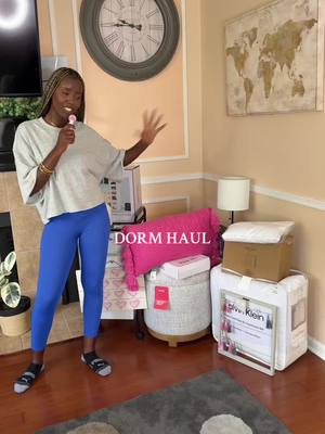 A young woman stands in a living room holding a microphone and gesturing, surrounded by boxes and dorm room supplies.