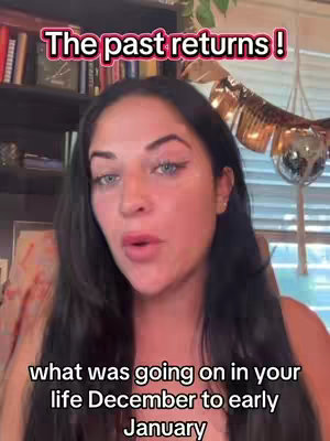 A woman with long black hair and fair skin is centered in a selfie-style shot. She appears to be talking. Overlaid at the top in a pink rectangle is the text "The past returns!". At the bottom is the text "what was going on in your life December to early January". The background shows a blurred bookshelf and a decorative item.