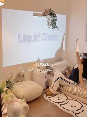 The cover depicts a room bathed in soft, warm light, a woman sits on a bed. The wall behind is used as the projection screen. A phrase "Liquid Glass" is projected, the room is decorated with cozy elements, including a floor rug with a cloud-like pattern. The scene radiates a comfortable, relaxed atmosphere.