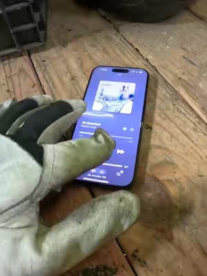 A person's gloved hand holds a smartphone displaying a music app interface. The phone is angled toward the camera, showcasing the screen against a backdrop of wooden planks. The gloved hand features an interesting texture, and the phone lights are bright.